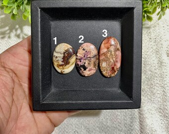 May include: Three oval-shaped gemstones with unique patterns and colors, displayed in a black square tray. Gemstone 1 is cream and brown, gemstone 2 is pink and black, and gemstone 3 is red and brown. The gemstones are numbered 1, 2, and 3.