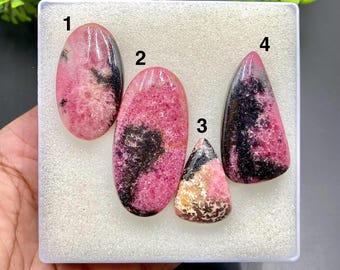 May include: Four polished rhodochrosite gemstones in varying shapes. The stones display a mix of pink and black hues, with some having a marbled appearance. The gemstones are displayed in a white plastic container.