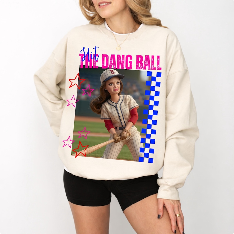 May include: Cream-colored sweatshirt featuring a graphic of a baseball player doll with the text "SHIT THE DANG BALL" in pink and blue. The design includes stars and a checkered pattern.