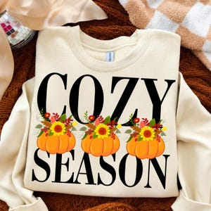 Könnte beinhalten: Ein cremefarbenes Sweatshirt mit Rundhalsausschnitt und dem schwarzen Schriftzug COZY SEASON in einer fetten Serifenschrift. Zwischen den beiden Wörtern befinden sich drei orangefarbene Kürbisse, die mit Sonnenblumen und Herbstblättern verziert sind.