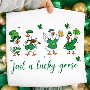 Puede incluir: Gráfico blanco del Día de San Patricio con cuatro gansos de dibujos animados. Cada ganso viste de verde y blanco, con sombreros y accesorios. El texto "just a lucky goose" está escrito en verde debajo.