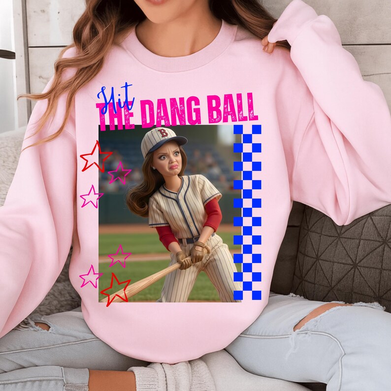 May include: Pink sweatshirt with a graphic of a baseball player and the text "Hit the Dang Ball." The graphic includes stars and a blue and white checkered pattern. The baseball player is wearing a baseball uniform and a baseball cap.