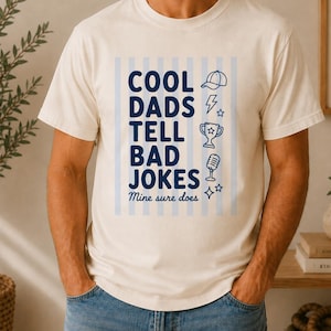 May include: A cream-colored short-sleeve t-shirt featuring a blue graphic with vertical stripes and the text "COOL DADS TELL BAD JOKES Mine sure does". The design includes small icons of a baseball cap, lightning bolt, trophy, and microphone.
