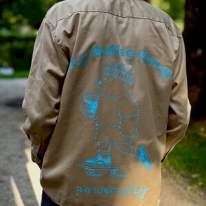 Könnte beinhalten: Beige Hemd mit blauem Aufdruck auf dem Rücken. Der Aufdruck zeigt einen Bären mit Mütze, der einen Kaffeebecher hält und skateboardet. Der Text "My Coffee Comp" und "powered by" sind ebenfalls blau.