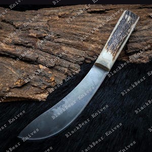 May include: A fixed-blade knife featuring a curved, dark steel blade and a textured, light-colored antler handle. The knife is positioned diagonally across the frame, showcasing the contrast between the smooth metal and the organic, rough surface of the handle.
