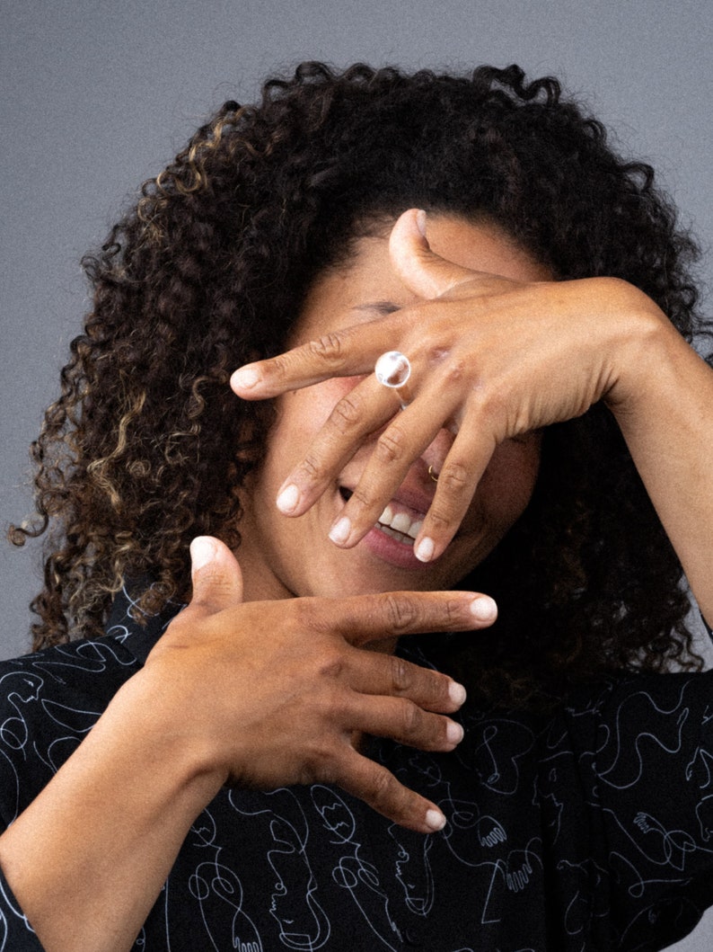 May include: A woman with curly hair is smiling and covering her face with her hands. She is wearing a black shirt with a white abstract pattern and a silver ring on her finger.