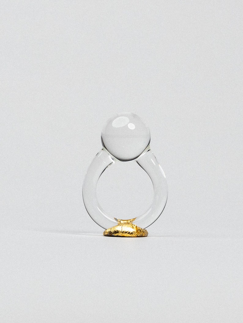May include: A clear glass ring with a round, clear glass top and a gold base.