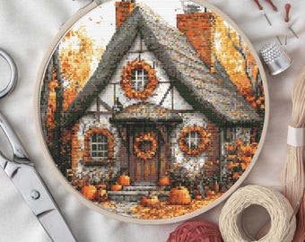 May include: A completed cross-stitch project of a cottage scene, set in autumn. The cottage has a gray roof, brick chimney, and orange wreaths. Pumpkins and fall foliage surround the house. The cross-stitch is in a wooden embroidery hoop, with scissors, thread, and pins nearby.