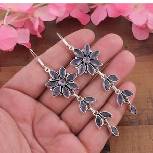 Può includere: Un paio di lunghi orecchini pendenti in argento con un design a forma di fiore, caratterizzato da pietre viola taglio marquise come petali e una piccola pietra viola rotonda al centro. Da ogni fiore pende una catena verticale di tre foglie viola.