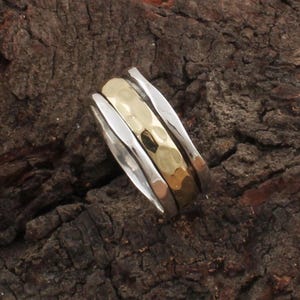 Puede incluir: Un anillo de tres bandas con una banda central dorada martillada, flanqueada por dos bandas plateadas. El anillo se presenta sobre una superficie de madera oscura y texturizada. El diseño es moderno y minimalista.