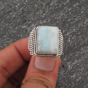 May include: A silver ring with a rectangular, light blue gemstone. The ring has a textured, beaded border and a twisted rope-like design around the gemstone. The ring is held in a hand.