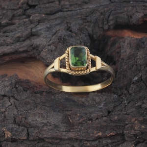 May include: A gold ring featuring a rectangular green gemstone set in a twisted rope-style metal frame. The band has a split design where it connects to the central setting, showcasing a polished metallic finish and a vibrant green stone.