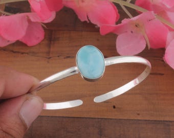 May include: A silver cuff bracelet featuring a single, smooth, oval-shaped light blue Larimar gemstone set in a polished metal bezel. The bracelet has an open-ended design with a slender, rounded band that tapers slightly towards the open ends.