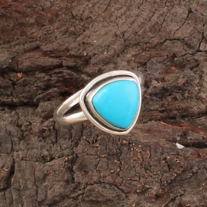 Pode incluir: Um anel de prata com uma pedra turquesa triangular e brilhante. A pedra é engastada em uma moldura de prata, criando um contraste marcante. O anel tem um design simples e elegante.