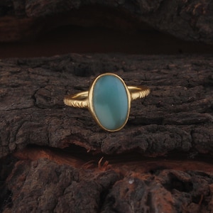May include: A gold ring featuring a smooth, oval-shaped, light blue-green cabochon gemstone set in a bezel. The ring band has a textured, etched surface pattern along the sides, providing a distinct contrast to the polished finish of the stone.
