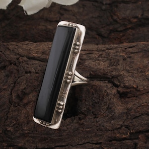 May include: A rectangular silver ring with a large, polished black stone. The ring has a decorative border with small, raised details. The ring is set against a dark, textured background.