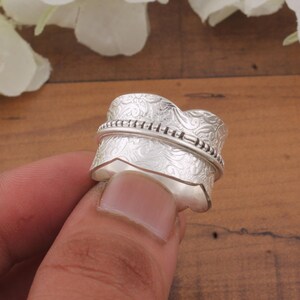 Puede incluir: Un anillo plateado con un intrincado diseño floral y una banda giratoria. El anillo está sostenido por un dedo, mostrando su diseño detallado. La banda tiene una textura de cuentas. Joyería.