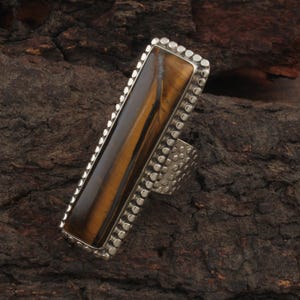 May include: A rectangular ring featuring a tiger's eye stone with brown and gold stripes. The stone is set in a silver-colored metal with a dotted border and a textured band. The ring is displayed on a dark brown, textured surface.