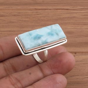 Könnte beinhalten: Rechteckiger Silberring mit einem großen, hellblauen Edelstein mit weißer Marmorierung. Der Edelstein ist in einer doppelten Silberfassung gefasst. Der Ring wird auf einer Holzoberfläche präsentiert.