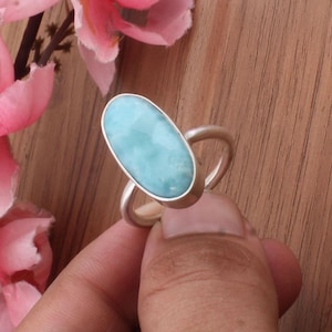 Könnte beinhalten: Ein Silberring mit einem großen, ovalen, hellblauen Edelstein. Der Edelstein ist in einer silbernen Lünette gefasst und hat eine glatte, polierte Oberfläche. Das Band des Rings ist schlicht und abgerundet. Der Hintergrund umfasst rosa Blumen und eine Holzoberfläche.