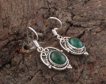 Può includere: Orecchini argentati con pietre preziose ovali verde smeraldo. Gli orecchini hanno un bordo decorativo in stile filigrana e una chiusura a gancio. Lo sfondo è una superficie marrone strutturata. Misurano circa 2 cm.