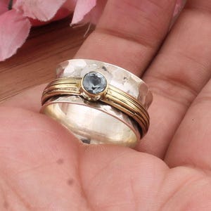 Puede incluir: Un anillo giratorio plateado y dorado con una piedra preciosa azul claro. El anillo presenta una textura martillada en la banda plateada y una banda dorada alrededor del centro. La piedra está engastada en un bisel dorado.