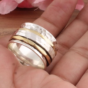 May include: A silver ring with a hammered texture and two gold bands. The spinner ring has gold bands that rotate around the silver band. The ring is displayed on a person's hand.