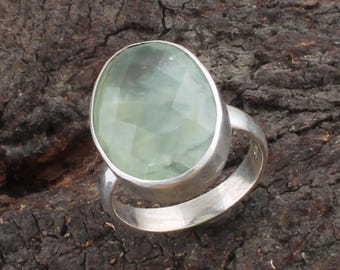 May include: A silver ring featuring a large, oval-cut, pale green gemstone. The gemstone has a faceted surface, catching the light. The ring is set on a dark, textured background, highlighting the jewelry.