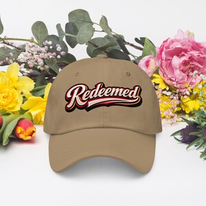 Puede incluir: Una gorra de béisbol de color tostado con la palabra Redeemed en una fuente cursiva estilizada de varias capas con contornos negros, blancos y rojos. La gorra tiene una corona estructurada, una visera curva y un botón en la parte superior.