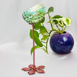 以下が含まれることがあります： 銅色の茎とベースが付いた装飾的な貝殻プランターで、鮮やかな緑とターコイズの貝殻を保持しています。小さな丸い紫色の花瓶に植物が入っており、貝殻プランターの隣にあります。