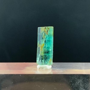 May include: A vibrant, translucent tourmaline crystal with shades of teal, yellow, and clear. The gemstone is vertically oriented and rests on a small, white base. The crystal is displayed on a reflective, rectangular surface against a dark background.