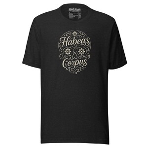 Puede incluir: Camiseta gris oscuro con un diseño de calavera color crema con detalles florales y las palabras "Habeas Corpus". El diseño está centrado en la parte delantera. La camiseta tiene cuello redondo y mangas cortas.