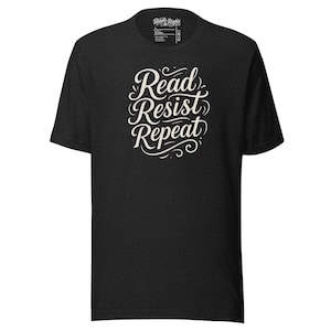 May include: A dark gray t-shirt with the words "Read Resist Repeat" in a white, cursive font. The shirt has a crew neck and short sleeves. The text is the focal point of the design.