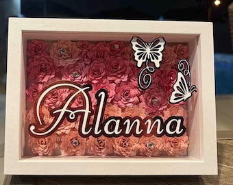 May include: A white framed shadow box filled with pink and peach paper roses. The name "Alanna" is displayed in a cursive font, with two butterfly cutouts. Rhinestones are placed in the center of some of the roses.