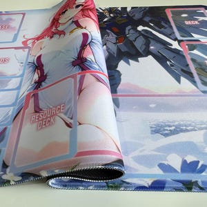 Puede incluir: Alfombrilla de juego rectangular enrollada, con ilustraciones de estilo anime. Un lado muestra un personaje femenino en blanco y rosa, el otro un robot. La alfombrilla tiene zonas de juego etiquetadas como "BASE", "SHIELDS" y "RESOURCE DECK".