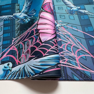 Puede incluir: Una alfombrilla de rat&oacute;n enrollada con un dise&ntilde;o de estilo c&oacute;mic. La obra de arte representa un paisaje urbano con palomas volando, una telara&ntilde;a rosa y una figura con un disfraz rosa y negro. La paleta de colores incluye tonos de azul, rosa y blanco.