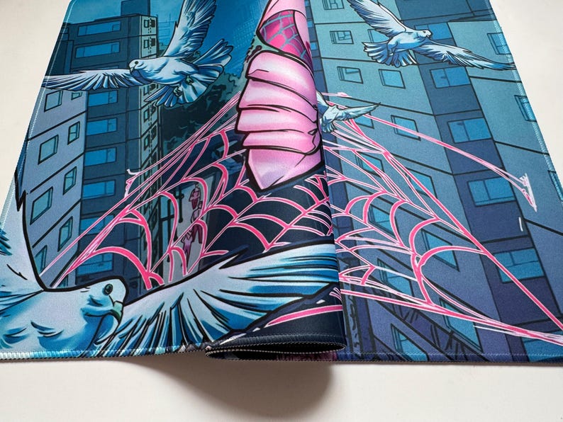 Puede incluir: Una alfombrilla de rat&oacute;n enrollada con un dise&ntilde;o de estilo c&oacute;mic. La obra de arte representa un paisaje urbano con palomas volando, una telara&ntilde;a rosa y una figura con un disfraz rosa y negro. La paleta de colores incluye tonos de azul, rosa y blanco.