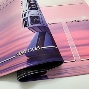 Puede incluir: Una alfombrilla de juego rosa y morada con una imagen estilizada de un robot y la palabra "RECURSOS". La alfombrilla tiene &aacute;reas designadas para el juego, con las palabras "DISCARD PILE" visibles. La alfombrilla est&aacute; enrollada, revelando un borde cosido negro.