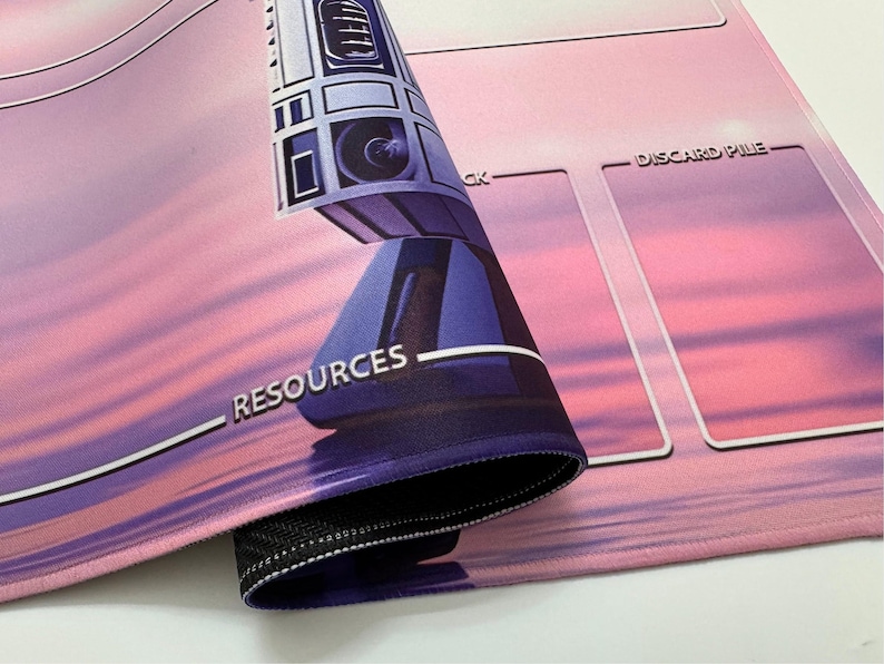 Puede incluir: Una alfombrilla de juego rosa y morada con una imagen estilizada de un robot y la palabra "RECURSOS". La alfombrilla tiene &aacute;reas designadas para el juego, con las palabras "DISCARD PILE" visibles. La alfombrilla est&aacute; enrollada, revelando un borde cosido negro.