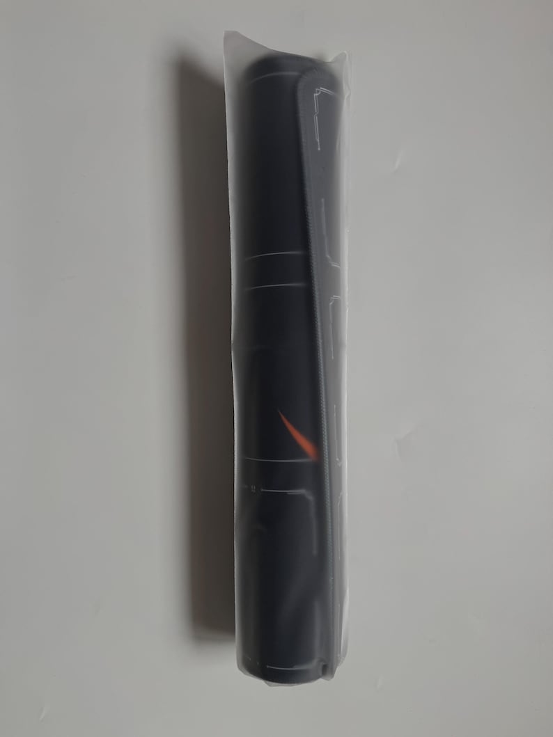 Op de afbeelding: Een opgerolde zwarte mat, verpakt in doorzichtig plastic. De mat heeft witte lijnen en een oranje ontwerp. Dit is waarschijnlijk een yogamat of een vergelijkbaar fitnessaccessoire.