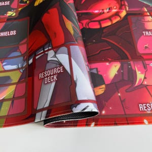 Puede incluir: Alfombrilla de juego roja y negra con una ilustraci&oacute;n detallada de un robot. La alfombrilla presenta secciones etiquetadas para "Base", "Shields", "Resource Deck" y "Trash". Los bordes est&aacute;n cosidos con hilo negro.