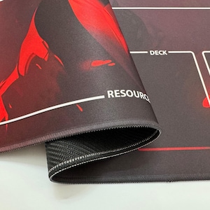 Puede incluir: Alfombrilla de juego con un dise&ntilde;o rojo y negro. La alfombrilla presenta &aacute;reas etiquetadas como "RESOURCE", "DECK" y "DISCARD". Los bordes est&aacute;n cosidos y la parte inferior tiene una superficie texturizada antideslizante.