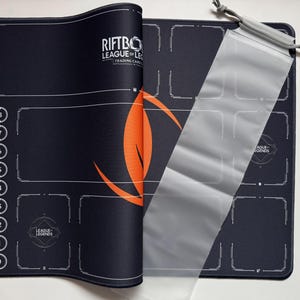 Op de afbeelding: Zwarte en oranje Riftbo League of Legends speelmat voor trading card games met een doorzichtige kaarthoes. De speelmat heeft een ontwerp met genummerde gebieden en het League of Legends logo.
