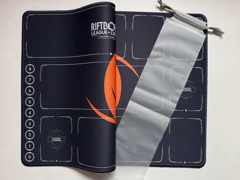 Op de afbeelding: Zwarte en oranje Riftbo League of Legends speelmat voor trading card games met een doorzichtige kaarthoes. De speelmat heeft een ontwerp met genummerde gebieden en het League of Legends logo.