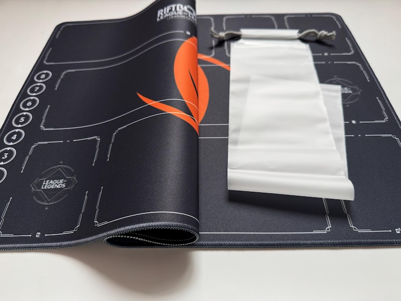Puede incluir: Alfombrilla de rat&oacute;n para juegos negra con gr&aacute;ficos blancos y naranjas, incluido el texto "RIFTTO", "LEAGUE LEGENDS". Se incluye una bolsa con cord&oacute;n blanca. La alfombrilla tiene un borde cosido y un dise&ntilde;o detallado.