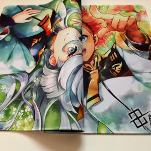 Op de afbeelding: Kleurrijke muismat met anime-stijl illustraties van twee personages. Het ontwerp bevat groene, witte en oranje tinten, met de tekst "GUNDAM CARD GAME" rechtsonder. Het kunstwerk is gedetailleerd.