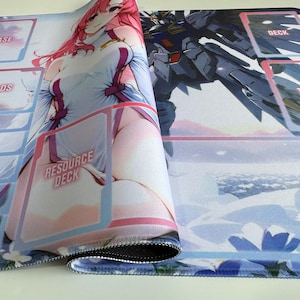 Op de afbeelding: Opgerolde speelmat met illustraties in anime-stijl. Het ontwerp bevat een vrouwelijk personage met roze haar en een robot. Tekst op de mat is onder meer "BASE", "SHIELDS", "RESOURCE DECK", "DECK" en "TRASH". De mat heeft een blauwe bloemenrand.
