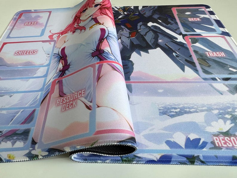 Op de afbeelding: Opgerolde speelmat met illustraties in anime-stijl. Het ontwerp bevat een vrouwelijk personage met roze haar en een robot. Tekst op de mat is onder meer "BASE", "SHIELDS", "RESOURCE DECK", "DECK" en "TRASH". De mat heeft een blauwe bloemenrand.
