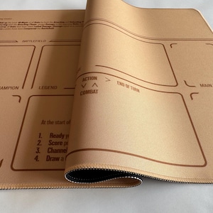 Op de afbeelding: Beige gamingmat met bruine contouren en tekst. De mat is opgerold en onthult een zwarte onderkant. De tekst bevat "BATTLEFIELD", "CHAMPION", "LEGEND" en "MAIN DECK". Er staan ook instructies op.