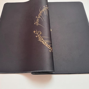 Puede incluir: Una alfombrilla de rat&oacute;n rectangular negra con un dise&ntilde;o dorado de escritura &eacute;lfica. La alfombrilla est&aacute; parcialmente enrollada, revelando la parte inferior. Los bordes est&aacute;n cosidos con hilo negro. El dise&ntilde;o es de El Se&ntilde;or de los Anillos.